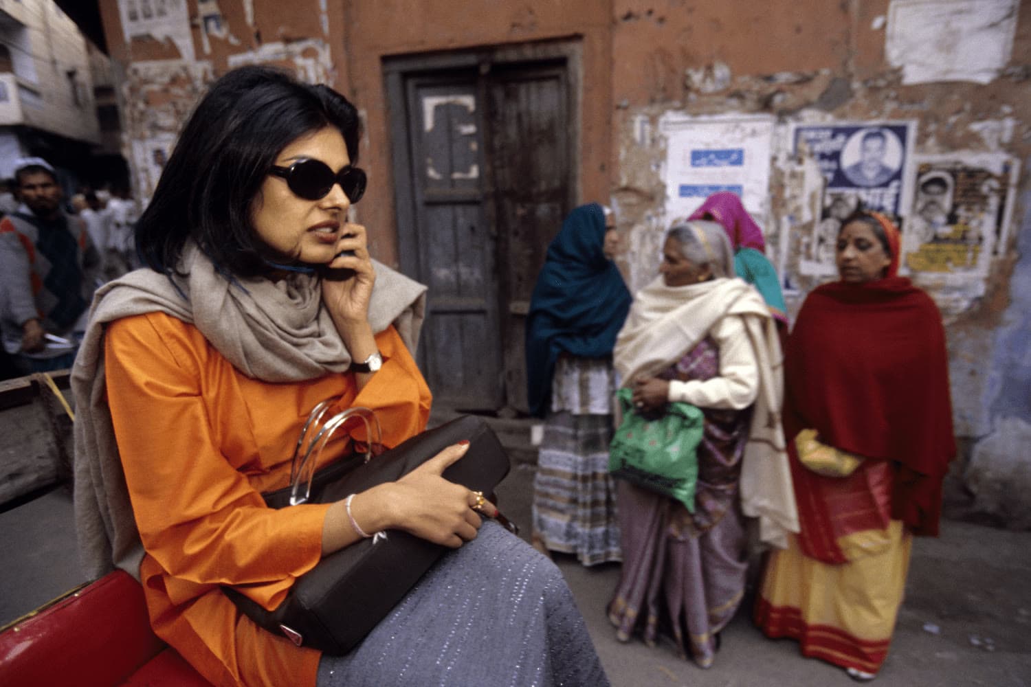 A woman in an orange kurta and sunglasses talks on a phone in an Indian street market while three women in saris stand near a weathered wall in the background.