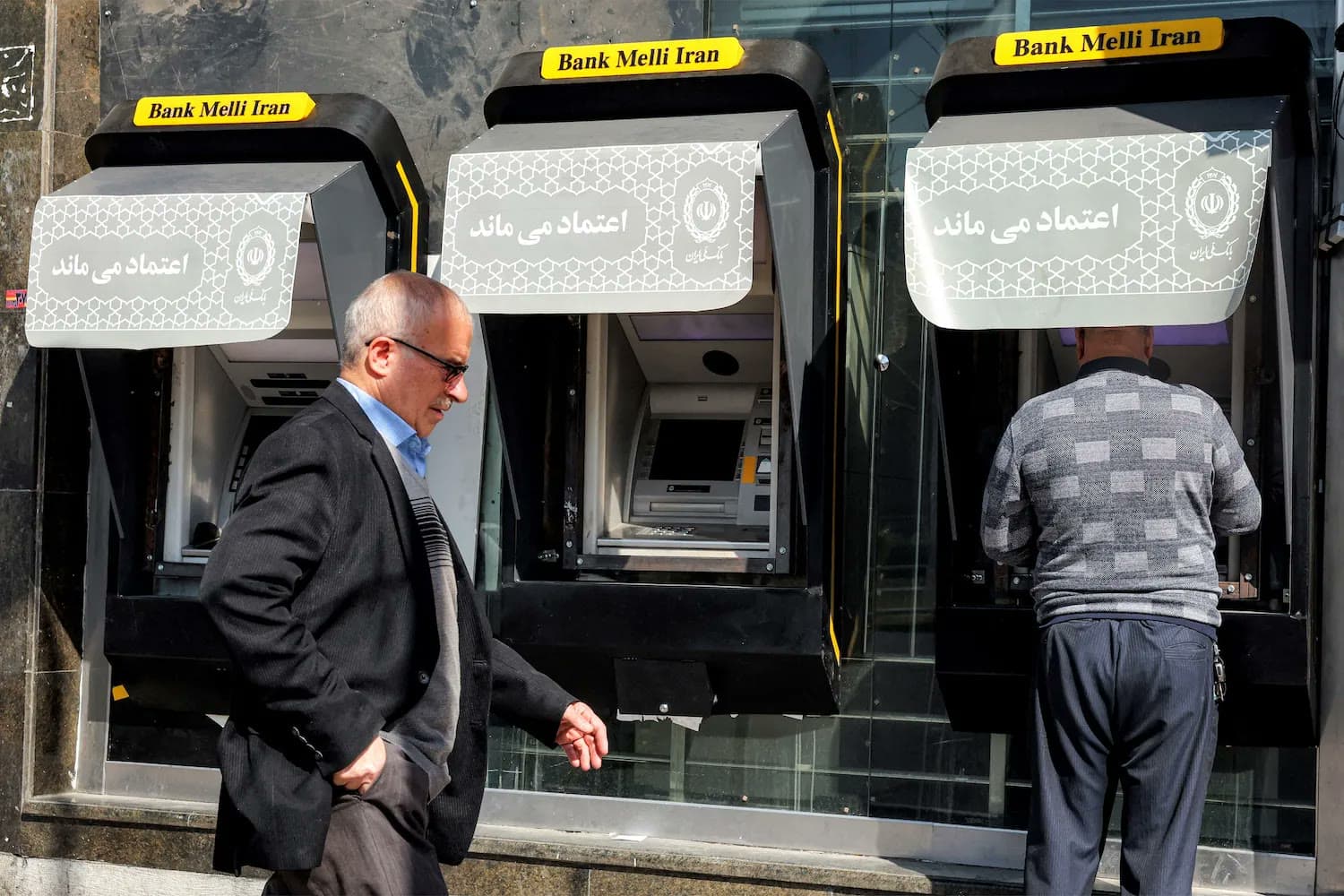Two men use ATMs outside Bank Melli Iran branches on a street in Tehran.