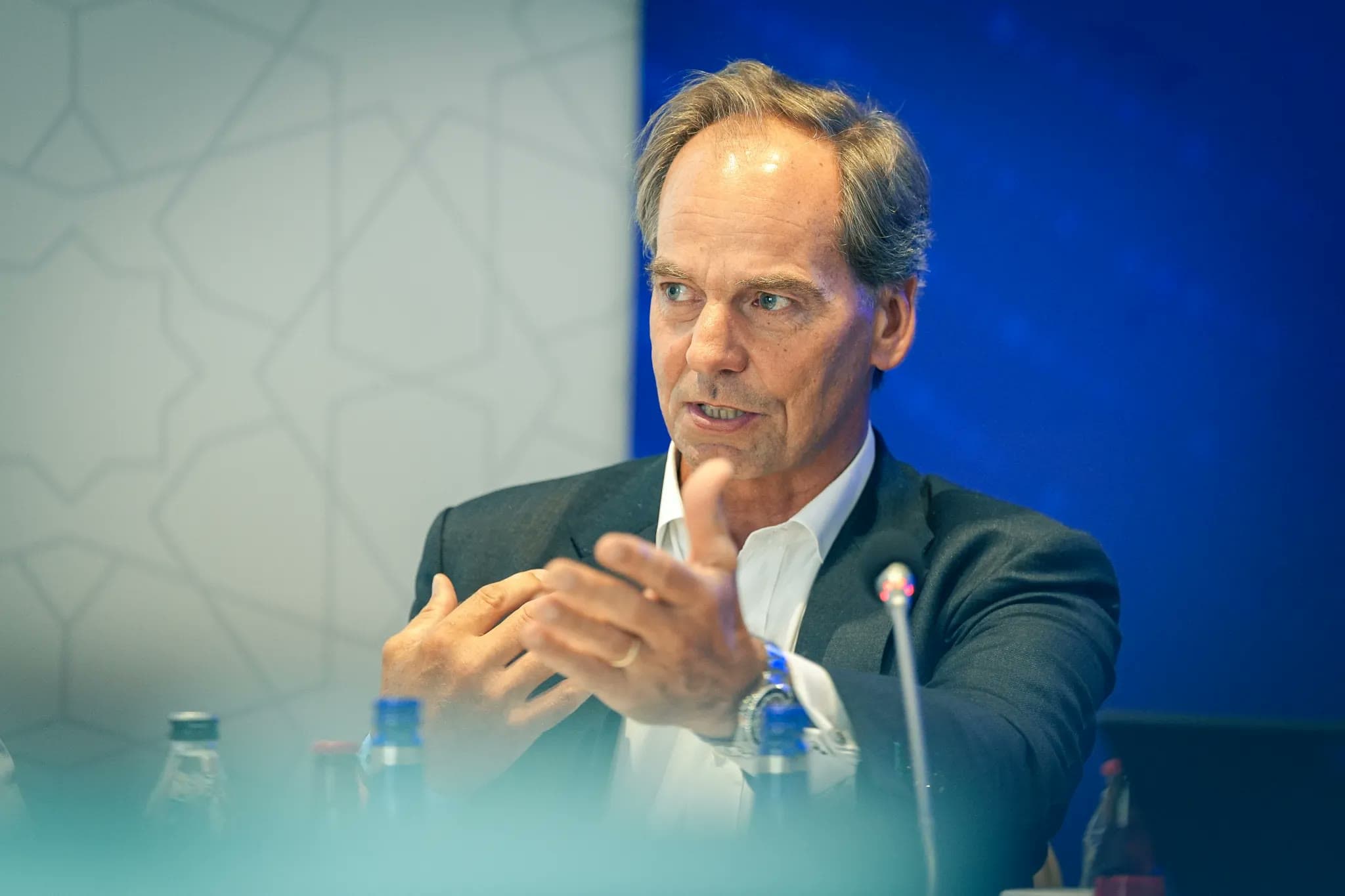 A speaker gestures while making a point during a panel discussion at a security conference, with a microphone and water bottles on the table in front of him.