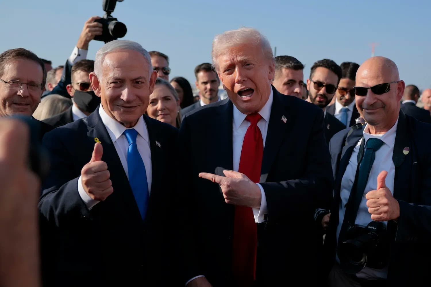 Benjamin Netanyahu and Donald Trump stand together giving thumbs-up and pointing gestures, surrounded by aides and photographers.