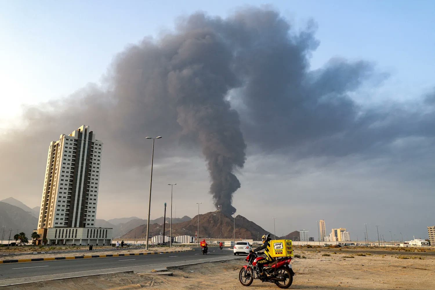 A large plume of black smoke rises over a coastal UAE city as a delivery rider on a motorbike passes in the foreground.