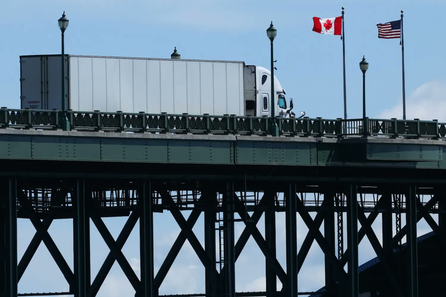 A semi truck crosses a steel bridge flying the Canadian and American flags at a U.S.-Canada border crossing.